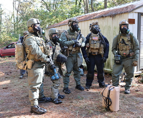 Group of people in tactical gear with gas masks outdoors near a building.
