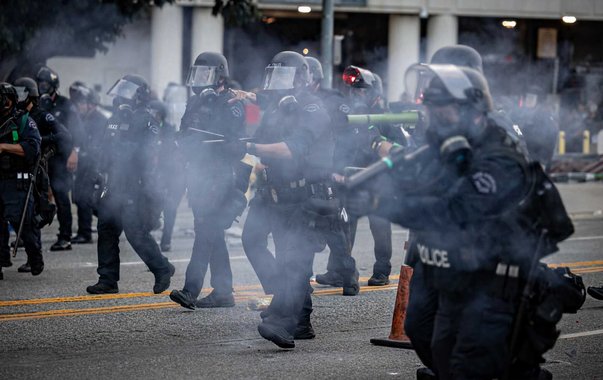 Police officers in riot gear during a protest with smoke and tear gas.