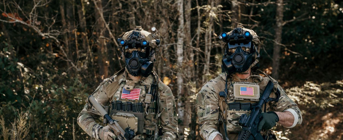 Two individuals in tactical gear with American flags on their vests, standing in a forested area.