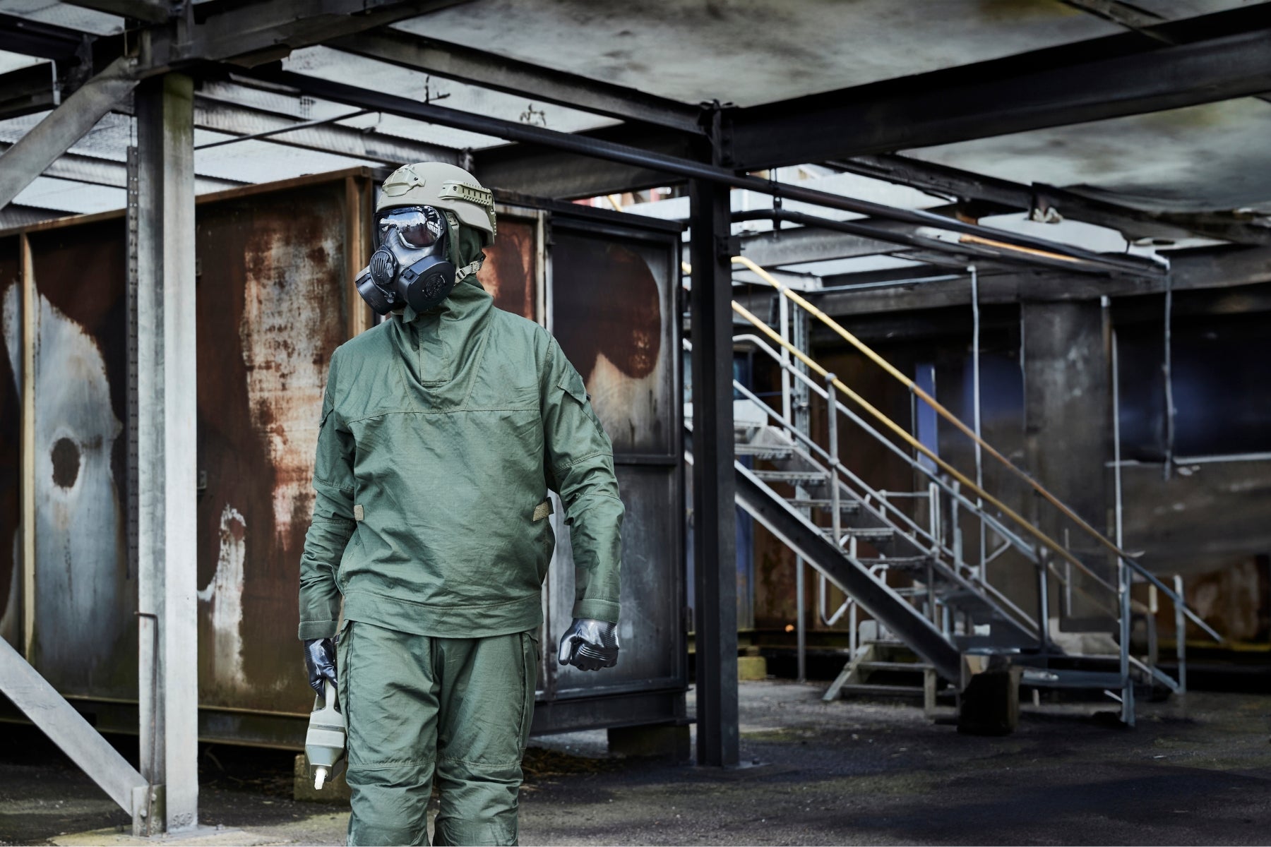 A person wearing a green tactical hazmat suit, helmet, and full-face respirator stands in an industrial outdoor setting.