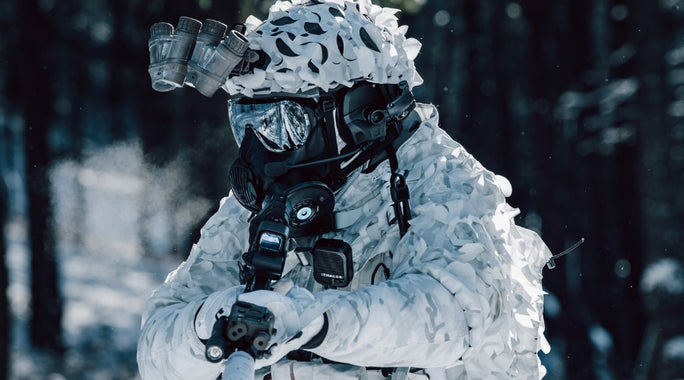 Person in full winter camouflage gear, gas mask, and a rifle in a snowy forest setting